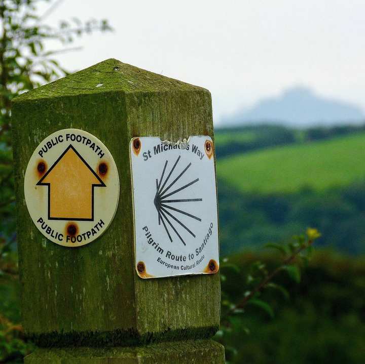 A waymark of Saint Michael's Way, the shell in a white circle, with a yellow footpath arrow, and Saint Michael's Mount beyond.