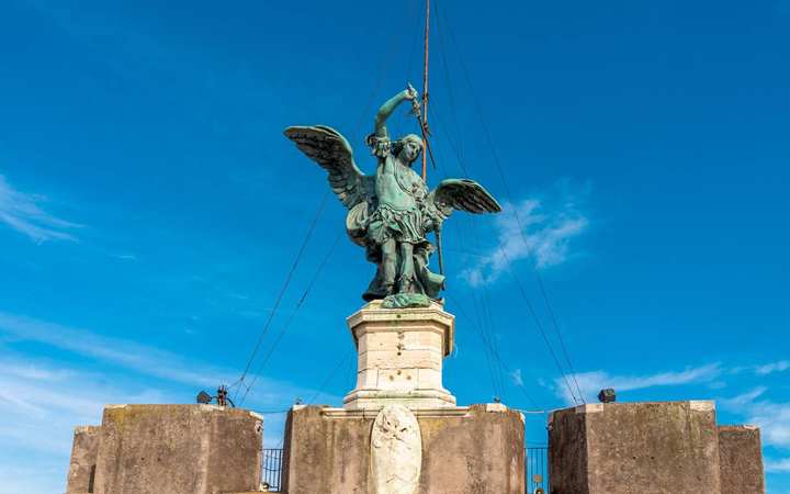 Michael over the plague — Hadrian’s Mausoleum, Rome. Looking up at a bronze archangel with spread wings on a stone pedestal, sword raised, against a clear sky. The statue stands on the roof of the round fortress in Rome named for this vision.