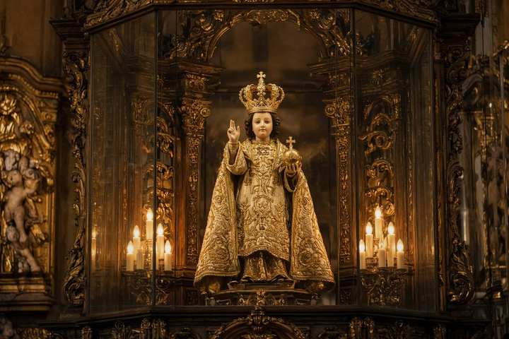 The Infant Jesus of Prague — Our Lady Victorious, Prague. The Infant of Prague in a white embroidered robe and gold crown, the globe in his left hand, two fingers raised in blessing.
