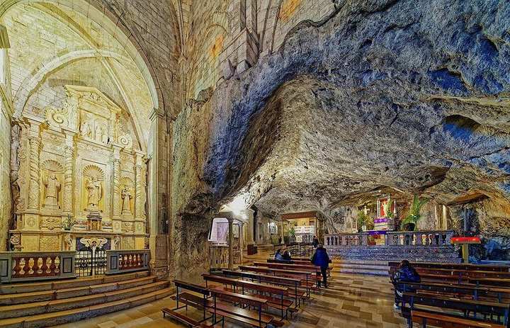 The cave of Saint Michael — Monte Sant’Angelo, Gargano, Apulia. Interior of a cave used as a church: rough rock ceiling, wooden pews, a carved altar on the left, lamps, a few pilgrims. This is the shrine St. Michael is said to have consecrated himself.